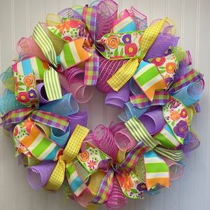 May include: A colorful spring wreath with pink, yellow, green, and blue mesh and ribbon. The ribbon features a floral pattern with white, orange, and pink flowers.