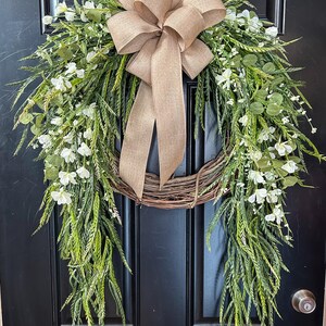 May include: A decorative wreath with a natural grapevine base, adorned with green foliage, white flowers, and a large burlap bow. The wreath is hanging on a black door, creating a welcoming aesthetic.