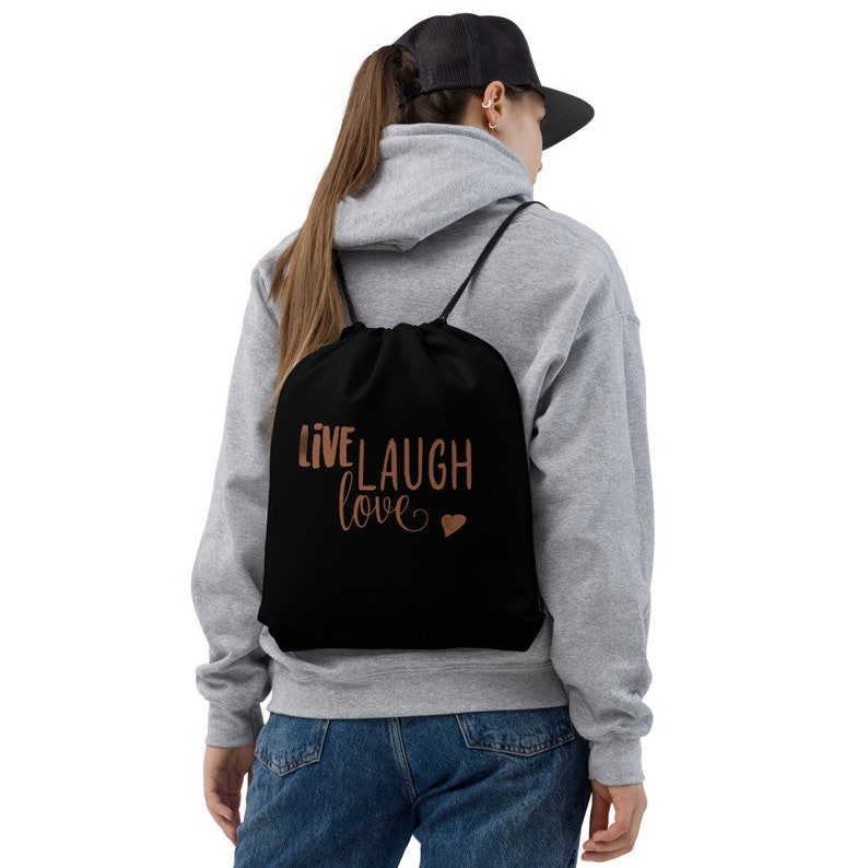 Peut inclure: Sac &agrave; cordon noir avec le texte "Live Laugh Love" en lettres couleur cuivre et un petit c&oelig;ur.