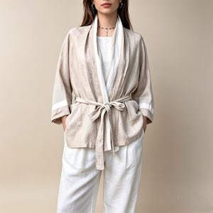 May include: A beige open-front jacket with a white inner lapel and a matching tie belt at the waist. The jacket features three-quarter length sleeves with white horizontal bands near the cuffs, worn over a white top and white trousers.