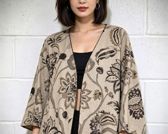 May include: A beige open-front jacket featuring a dark brown floral and botanical print. The garment has three-quarter length sleeves, a V-neckline with thin dark piping along the edges, and two front pockets integrated into the floral pattern.
