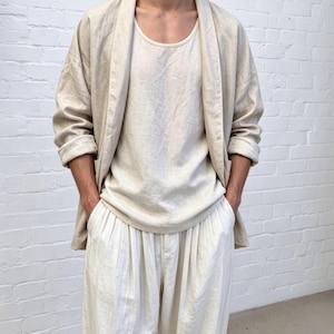 Puede incluir: Una chaqueta beige abierta con mangas largas, usada sobre una camiseta sin mangas beige a juego con cuello redondo y pantalones holgados de color blanco roto. El conjunto presenta una apariencia de tela texturizada y silueta relajada.