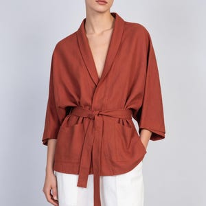 May include: A rust-colored wrap jacket featuring a shawl collar, wide three-quarter length sleeves, and two large front patch pockets. The garment is secured at the waist with a matching fabric tie belt, creating a structured yet relaxed silhouette.