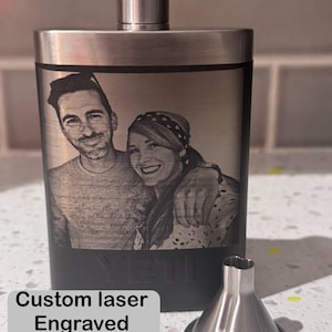May include: A black and silver YETI flask with a custom laser-engraved image of a couple. The flask has a silver cap and a matching silver funnel. The text "Custom laser Engraved YETI Flask" is in a white box.