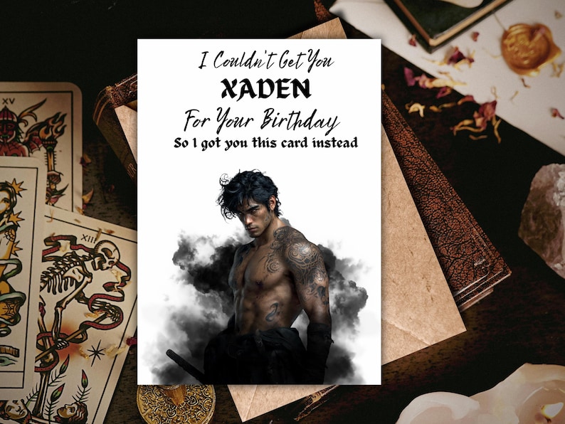May include: A greeting card with the text "I Couldn't Get You XADEN For Your Birthday So I got you this card instead." The card features an illustration of a shirtless man with tattoos and dark hair. Tarot cards and a book are in the background.