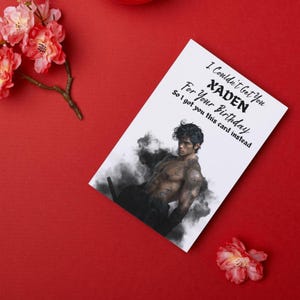 May include: A white greeting card with the text "I Couldn't Get You XADEN For Your Birthday So I got you this card instead." The card features an illustration of a muscular person. The card is on a red surface with pink flowers.