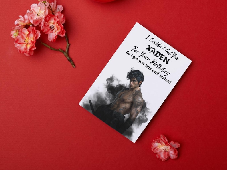 May include: A white greeting card with the text "I Couldn't Get You XADEN For Your Birthday So I got you this card instead." The card features an illustration of a muscular person. The card is on a red surface with pink flowers.