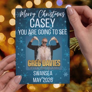 May include: A festive Christmas card with a blue backdrop and snowflake accents. The card features the text "Merry Christmas CASEY, YOU ARE GOING TO SEE GREG DAVIES, FULL FAT LEGEND, SWANSEA, MAY 2026." A photo of a man is in the center.