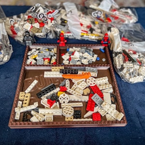 May include: A brown plastic tray filled with various small white, gray, and red Lego pieces. The tray is divided into sections and has a raised edge. There are also several bags of Lego pieces in the background.