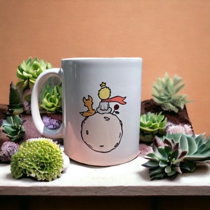 May include: White ceramic mug with a black and white illustration of The Little Prince sitting on an asteroid with a fox. The mug has a black handle and is surrounded by succulents.