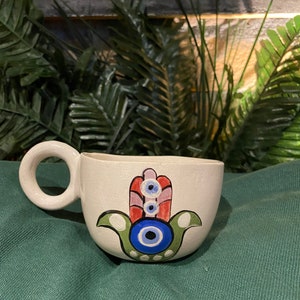 May include: White ceramic mug with a hand-painted hamsa design. The hamsa is green, red, and blue with a blue evil eye in the center.