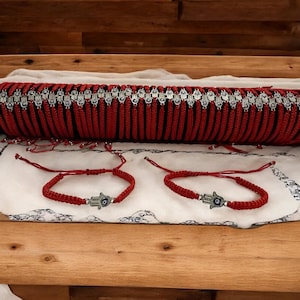 May include: Red woven bracelets with a Hamsa hand charm. The bracelets are displayed on a white cloth, with a larger red woven piece in the background. The Hamsa hand charm has a blue eye in the center.