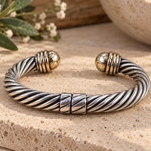 May include: A silver-toned cuff bracelet featuring a twisted cable design throughout the band. The bracelet has two polished gold-toned spherical end caps and a central rectangular hinge component that connects the two halves of the twisted metal.