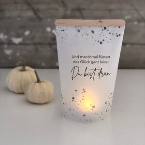 Puede incluir: Portavelas blanco con motas negras y parte superior de madera. El portavelas tiene el texto "Und manchmal flüstert das Glück ganz leise: Du bist dran" impreso en él.