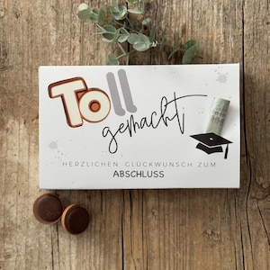 Puede incluir: Una caja de regalo blanca con las palabras "Toll gemacht" en letras naranjas y negras, junto con el texto "HERZLICHEN GLÜCKWUNSCH ZUM ABSCHLUSS". La caja está decorada con un birrete y un billete enrollado. Dos bombones en primer plano.