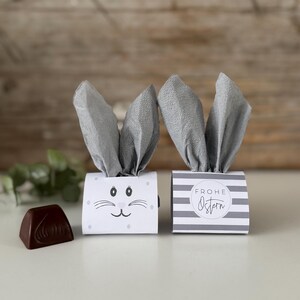 Könnte beinhalten: Zwei Oster-Geschenkboxen mit grauen Papier-Hasenohren. Eine Box zeigt ein Hasengesicht, die andere hat grau-weiße Streifen und den Schriftzug "Frohe Ostern". Eine Schokoladen-Süßigkeit liegt links.