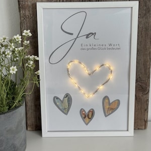 Puede incluir: Una obra de arte enmarcada con un borde blanco. La obra presenta la palabra "Ja" en cursiva, con texto debajo. Una cadena de luces en forma de corazón ilumina el centro. Tres imágenes en forma de corazón de moneda están debajo.