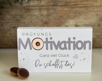 May include: White wooden block with the text "Prüfungs Motivation" in gray lettering. The word "Motivation" has a donut graphic with a white center and orange filling. The text "Ganz viel Glück" and "Du schaffst das!" are also printed on the block.