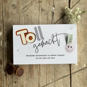 Può includere: Un biglietto di auguri bianco con le parole "Toll gemacht" in arancione e grigio, e un carattere scritto a mano. Il biglietto ha un adesivo con la faccina sorridente e il testo "Herzlichen Glückwunsch zu deinem Zeugnis! Ich bin stolz auf dich!" Due caramelle al cioccolato in basso.