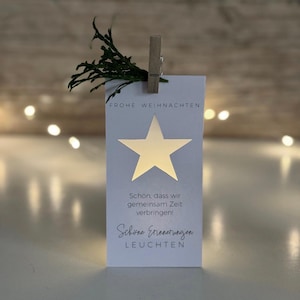 Könnte beinhalten: Eine weiße rechteckige Karte mit einem goldenen Stern-Ausschnitt und dem deutschen Text "FROHE WEIHNACHTEN" und anderem Text. Ein kleiner Zweig grünes Laub ist mit einer Holzklammer oben befestigt. Die Karte wird von hinten beleuchtet.