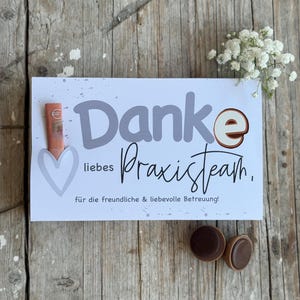 Op de afbeelding: Een witte kaart met de Duitse woorden "Danke liebes Praxisteam" in grijze en zwarte letters, met een hart en een klein boeket witte bloemen. Twee chocolade snoepjes liggen op het houten oppervlak.