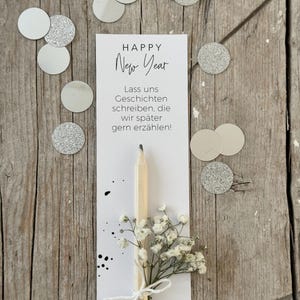 Op de afbeelding: Een witte kaart met de tekst "HAPPY New Year" en "Lass uns Geschichten schreiben, die wir später gern erzählen!" Een houten potlood met een klein boeket witte bloemen is aan de kaart bevestigd.
