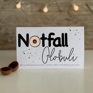 Pode incluir: Cartão branco com texto preto que diz "Notfall Globuli" e "Bei außergewöhnlichem Stress oder in Notfallsituationen". Há um pequeno círculo branco com um centro marrom no cartão.