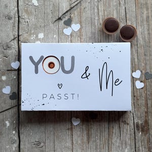 May include: A white card with black ink reads "YOU & Me" with a heart symbol and the word "PASST!"  The word "YOU" has a small circle with a brown center.  Two small chocolate candies are on a wooden surface.