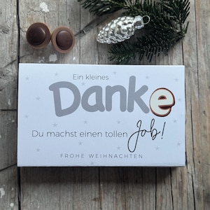 Peut inclure: Une boîte cadeau blanche avec du texte allemand disant "Danke" et "Du machst einen tollen Job !" avec deux chocolats et une décoration de pomme de pin argentée sur une surface en bois.