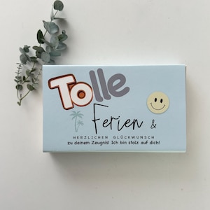 May include: A light blue rectangular card featuring the text "Tolle Ferien &" in a mix of bold and script fonts, a small palm tree icon, a yellow smiley face sticker, and the text "HERZLICHEN GLÜCKWUNSCH zu deinem Zeugnis! Ich bin stolz auf dich!"