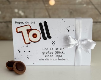 May include: White gift box with a white ribbon and a black and white design. The box has the text "Papa, du bist Toll" and "und es ist ein großes Glück, einen Papa wie dich zu haben!" on it. There are two chocolate candies in the foreground.
