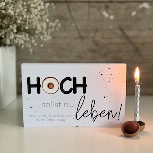 May include: White wooden sign with black text that reads "HOCH sollst du leben!" with a small donut graphic in the word "HOCH". The sign also has black splatter paint and the text "Herzlichen Glückwunsch zum Geburtstag!"  A silver birthday candle is lit in the background.