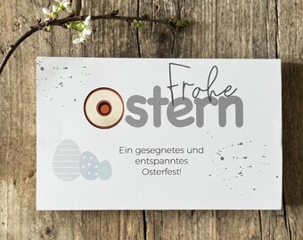 May include: A white card with the text "Frohe Ostern" in gray lettering. The card also has a small, round, wooden egg with a red center and the text "Ein gesegnetes und entspanntes Osterfest!" in gray lettering.