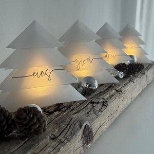 Pode incluir: Decoração de Natal com quatro lanternas em forma de árvore de papel branco, iluminadas por dentro. As lanternas trazem as inscrições « eins », « zwei », « drei » e « vier » e estão colocadas sobre uma base de madeira envelhecida, adornada com pinhas e bolas prateadas.