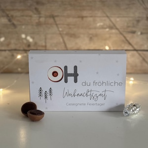 Peut inclure: Une boîte cadeau blanche avec le texte "OH du fröhliche Weihnachtszeit" et "Gesegnete Feiertage!" La boîte a un cercle rouge et blanc avec la lettre "H" au centre. Il y a trois petits sapins verts sur la boîte.