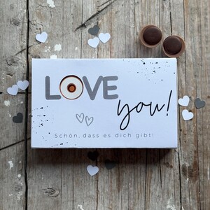 Könnte beinhalten: Eine weiße Karte mit dem Wort "LOVE" in grauen Buchstaben mit einem roten Kreis im "O". Die Karte sagt auch "you!" in schwarzer Schrift und "Schon. dass es dich gibt!" in grauem Text. Die Karte ist von weißen und grauen Papierherzen umgeben.