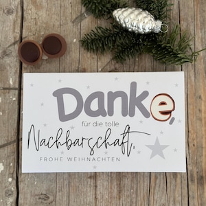 May include: A rectangular white card with the German word "Danke" in gray, and "Nachbarschaft" in black script. The card is decorated with gray stars and the words "Frohe Weihnachten". Two brown circular objects and a silver pinecone ornament are also present.