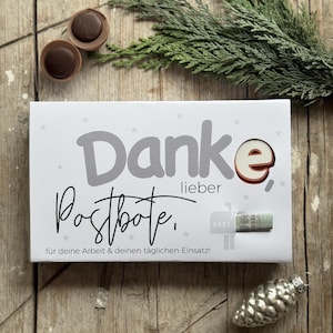 Puede incluir: Una caja de regalo blanca con el texto alemán "Danke lieber Postbote" y un pequeño gráfico de buzón. La caja está decorada con formas de estrella y un caramelo de chocolate. También hay una ramita de hoja perenne y un adorno plateado.