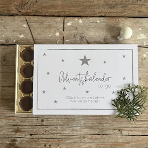 Peut inclure: Une boîte de calendrier de l'Avent rectangulaire avec un couvercle blanc orné d'étoiles grises et de texte. La boîte contient plusieurs chocolats. Le texte se lit "Adventskalender to go" et "Glück ist, einen Lehrer wie Sie zu haben!"