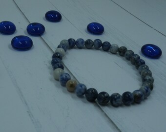 May include: A blue and white gemstone bracelet with round beads. The bracelet is on a white wooden surface with blue glass stones scattered around it.