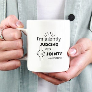 Puede incluir: Taza de cerámica blanca con un diseño negro que dice "Estoy juzgando silenciosamente tus articulaciones - Fisioterapeuta".