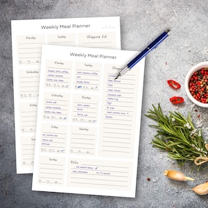Pode incluir: Duas folhas creme de Weekly Meal Planner com planos de refeições e listas de compras manuscritos. Uma caneta azul repousa sobre as folhas. Uma pequena tigela de grãos de pimenta, alecrim e dentes de alho está no fundo.