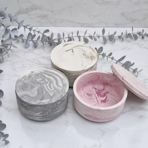 May include: Three round, decorative boxes with lids. The boxes are made of concrete and have a marbled design. One box is white with gold swirls, one is grey with white swirls, and one is pink with white swirls.