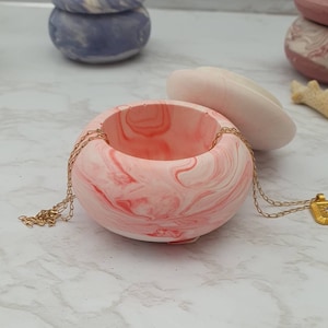 May include: A pink and white marbled ceramic trinket dish with a gold chain necklace and a gold pendant. The dish has a lid that is off to the side.
