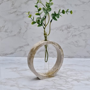 May include: A white and gold circular vase with a glass test tube in the centre. The vase is made of concrete and has a rough texture. A bouquet of small white flowers and green leaves is in the test tube.