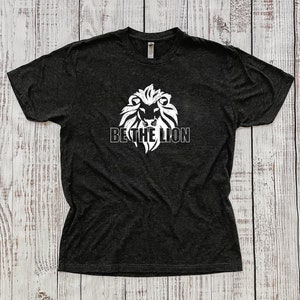 Peut inclure: Un t-shirt gris foncé avec un motif blanc représentant une tête de lion et le texte "BE THE LION".