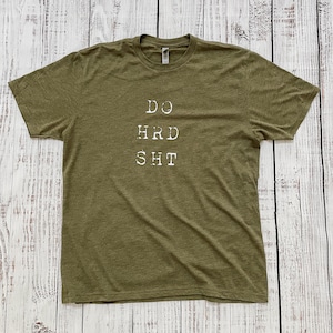 May include: Olive green t-shirt with the text "DO HRD SHT" printed in white. The shirt is made of a soft, textured fabric and is laid flat on a white wooden surface. The text is centered on the front of the shirt.