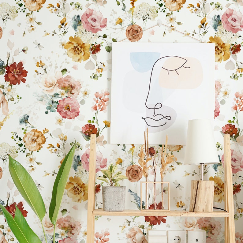 May include: A white wall with a floral wallpaper featuring pink, yellow, and red flowers. A wooden shelf with a white lamp, a potted plant, and a framed abstract line drawing of a face.