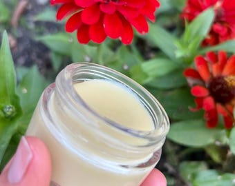 Può includere: Un piccolo barattolo di vetro trasparente riempito con un balsamo giallo chiaro. Il barattolo è tenuto davanti a fiori rossi vibranti con fogliame verde sullo sfondo. Il balsamo appare liscio e cremoso.