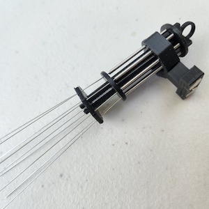 K&ouml;nnte beinhalten: Ein schwarzes Plastikwerkzeug mit mehreren d&uuml;nnen Metallnadeln, die sich von einem Ende erstrecken. Das Werkzeug hat eine Reihe von Federn und eine kleine Schlaufe am anderen Ende.